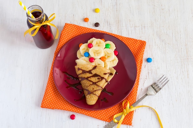 Children's breakfast or dessert - pancake with banana, chocolate topping and colorful candies. Making sweet food in the form of ice cream.