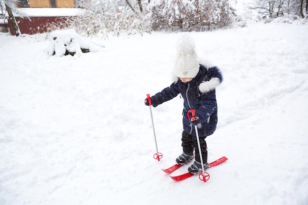 Children's feet in red plastic skis with sticks go through the snow from a slide-a winter sport, family entertainment in the open air. A little girl glides down the slope from an early age. Copy space