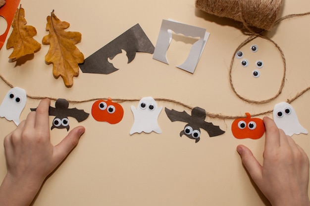 Children's hahomemade paper garland for Halloweennds next to a garland on a jute rope made of pumpkins, bats and ghosts with eyes on a beige background with paper scraps and autumn leaves