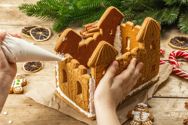 Children's hand decorate Gingerbread House by sweet icing. Handmade process, festive New Year decor. Traditional spices, wooden background, close up