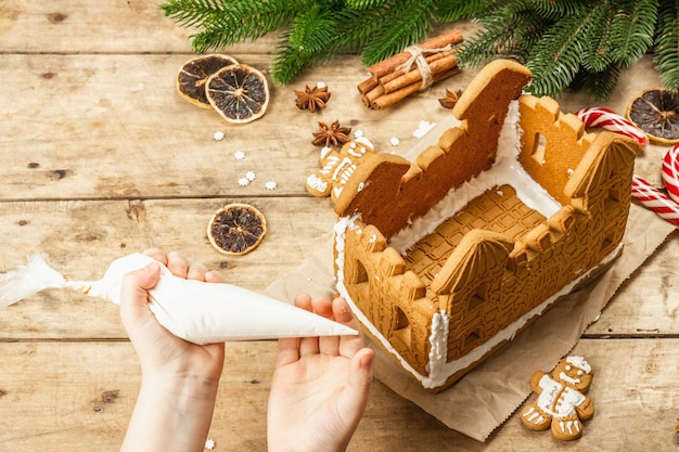 Children's hand decorate Gingerbread House by sweet icing. Handmade process, festive New Year decor. Traditional spices, wooden background, close up