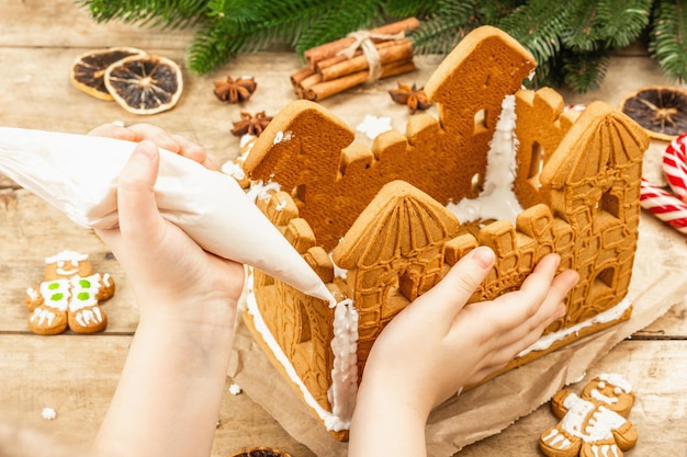 Children's hand decorate Gingerbread House by sweet icing. Handmade process, festive New Year decor. Traditional spices, wooden background, close up