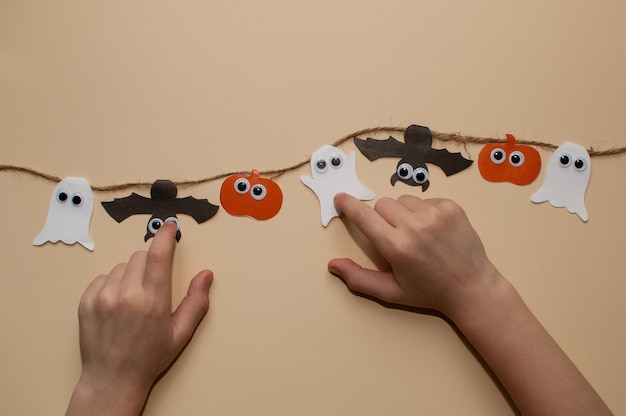 Children's hands hold a garland on a jute rope made of pumpkins bats and ghosts with eyes