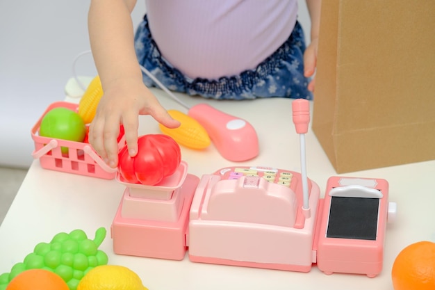 Children's hands weighs tomatoes on scales little girl plays in children's store at home with plastic toys toy cash register scanner