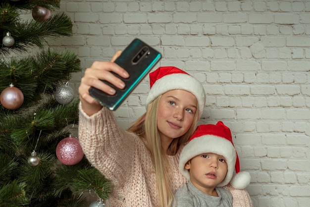 Children in Santa hats making selfie near Christmas tree at home, cute siblings in santa hats