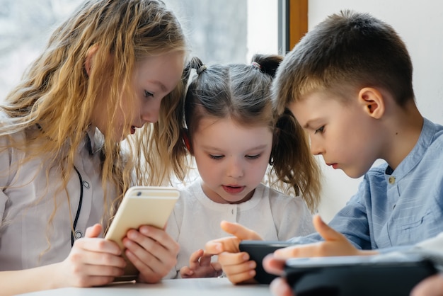 Children sit at a table in a cafe and play mobile phones together. Modern entertainment.