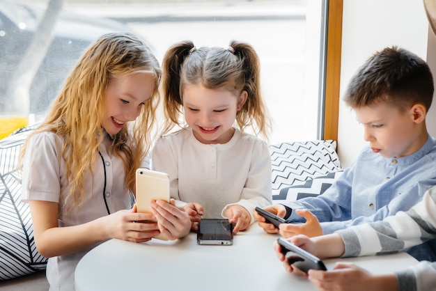 Children sit at a table in a cafe and play mobile phones together. Modern entertainment.