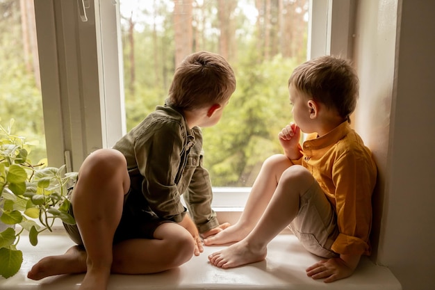 Children sitting on windowsill and waiting for someone comming Two brothers friends Cute preschool kids alone at home Boys are waiting for their mother or father Loneliness Busy parents