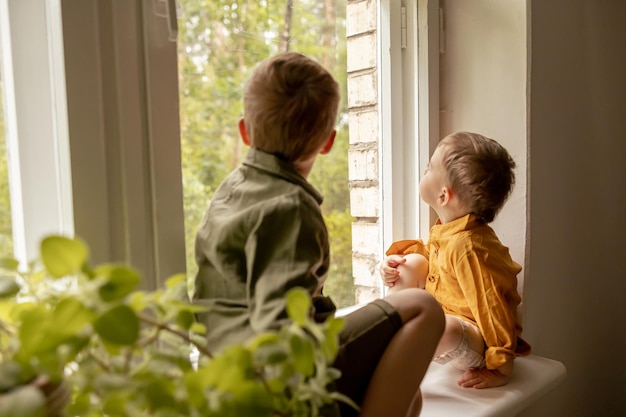 Children sitting on windowsill and waiting for someone comming Two brothers friends Cute preschool kids alone at home Boys are waiting for their mother or father Loneliness Busy parents