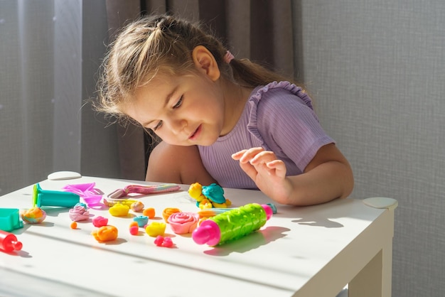 Children with plasticine little blonde girl enthusiastically plays with plasticine sculpts various colorful figurines from plasticine at home