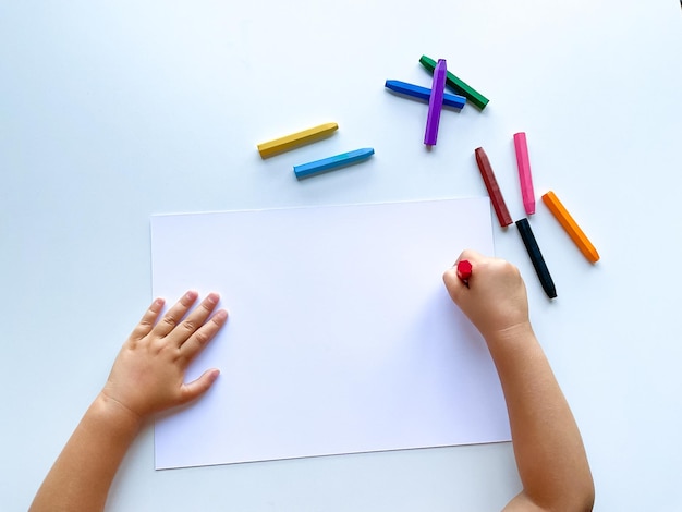 Childrens hands draw with colored wax crayons on a white sheet of paper Top view of a blank sheet