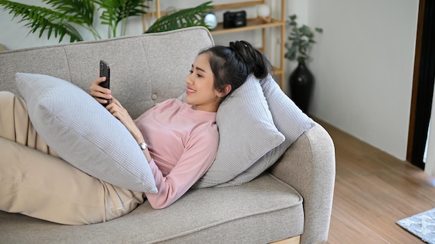 Chilling Asian female laying on sofa while scrolling on her social media on mobile phone