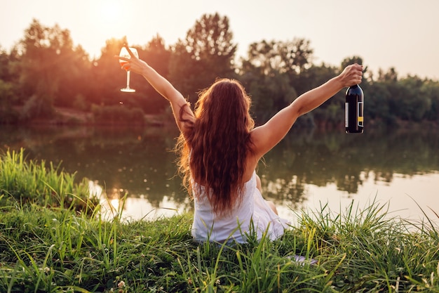 Photo chilling with wine. woman enjoying glass of wine on river bank at sunset raising arms and feeling free and happy.