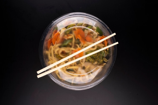 Chinese food delivery vegetarian yakisoba bowl top view black background with chopsticks and plastic lid