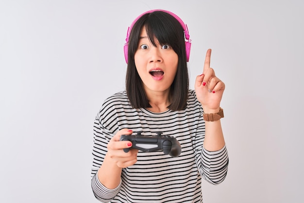 Chinese gamer woman playing video game using headphones over isolated white background surprised with an idea or question pointing finger with happy face number one