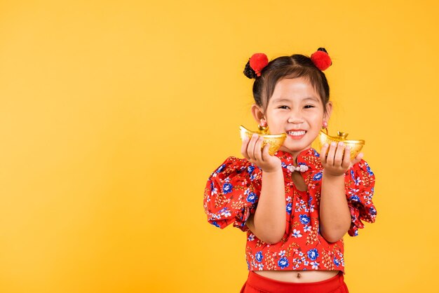 Chinese New Year Happy Asian Chinese little girl smile wearing red cheongsam holding gold ingot Portrait children in traditional dress hold golden bar studio short isolated on yellow background