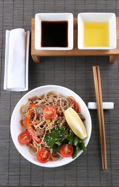 Chinese noodles with vegetables and roasted meat in bowl on napkin on wooden background