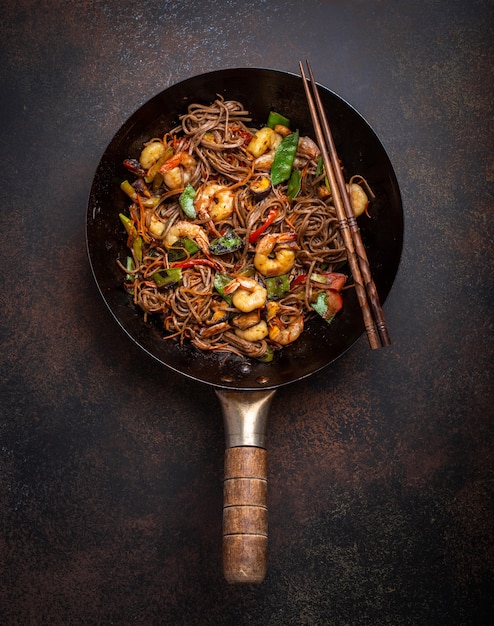 Chinese soba stir-fry noodles with shrimps, vegetables in old rustic wok pan served on concrete background, close up, top view. Traditional asian/thai dish, close up