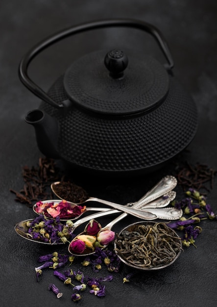 Chinese teapot with cup and spoons with various tea on black background Rose budsblue mallow flowersgreen and black loose tea