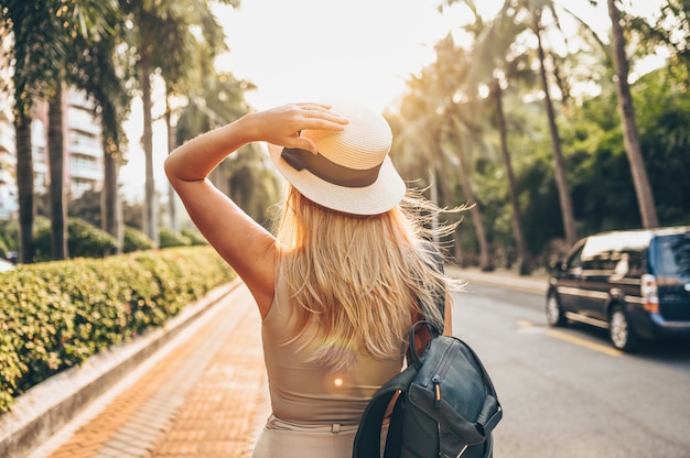 Chinese tourist walking in city streets on Asia vacation tourism. Caucasian beautiful woman in hat with backpack travel lifestyle concept. Walking street, green palms and highway in Sanya, Hainan.