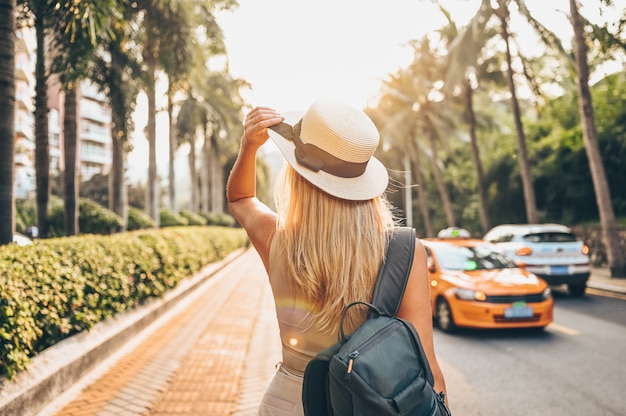 Chinese tourist walking in city streets on Asia vacation tourism. Caucasian beautiful woman in hat with backpack travel lifestyle concept. Walking street, green palms and highway in Sanya, Hainan.