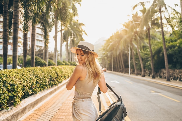 Chinese tourist walking in city streets on Asia vacation tourism. Caucasian beautiful woman in hat with backpack travel lifestyle concept. Walking street, green palms and highway in Sanya, Hainan.