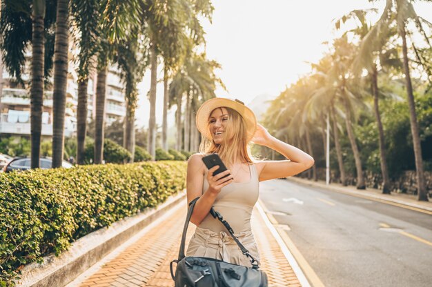 Chinese tourist walking in city streets on Asia vacation tourism. Caucasian beautiful woman with backpack, smartphone. Travel lifestyle concept. Street with green palms and highway in Sanya, Hainan.