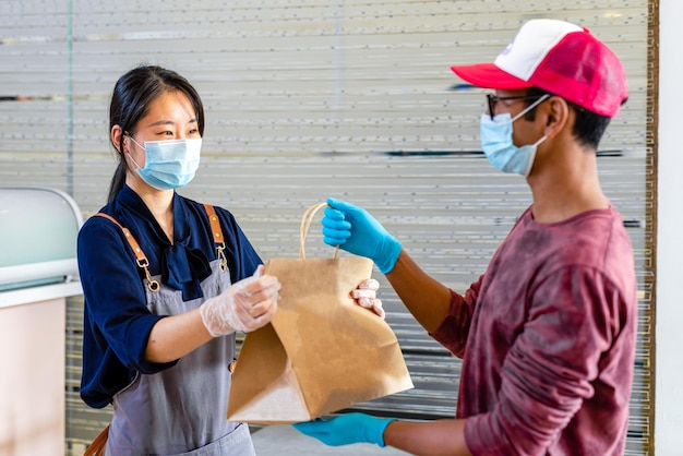 Chinese waitress with gloves and protective mask delivers to a courier the takeaway food in a paper bag protection against contagion at work the new normal concept in the restaurant business