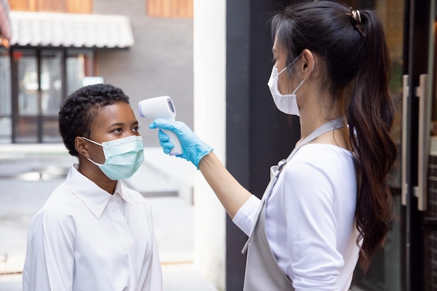 Chinese woman retail business staff or grocery shop keeper checking fever of black African customer by portable thermo scan device