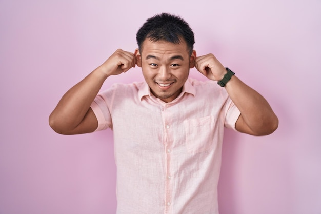 Chinese young man standing over pink background smiling pulling ears with fingers, funny gesture. audition problem