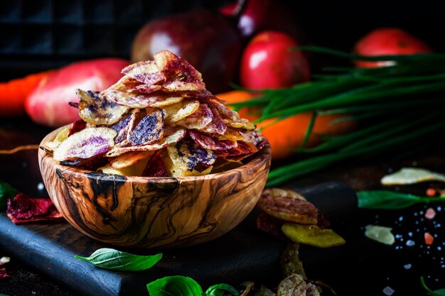 Chips from purple and red potatoes in wooden bowl on background of vegetables selective focus