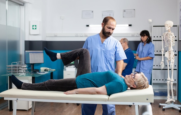 Chiropractor giving assistance to senior patient with knee injury for physical recovery. Woman doing leg exercise with help from medical assistant for osteopathic remedy and treatment.