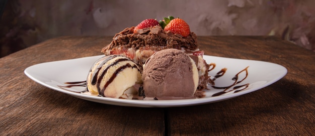 Chocolate cake, a tasty slice of chocolate cake with strawberries and ice cream balls on a white plate on rustic wood, abstract background, selective focus.