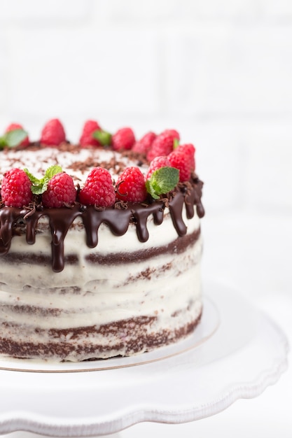 Chocolate cake with white cheese cream decorated ganache and raspberries on a white cake stand