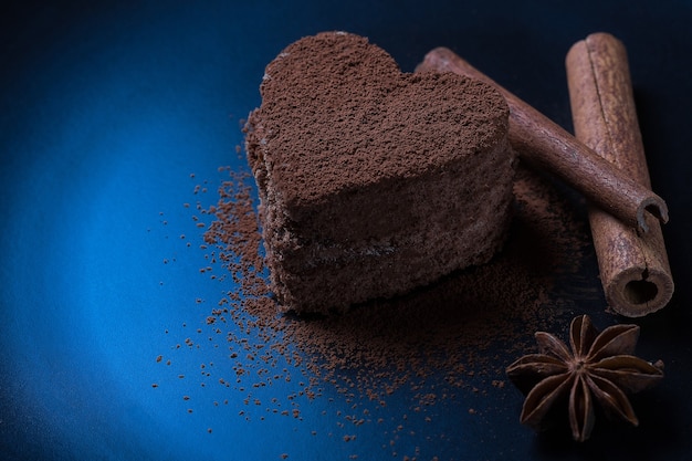 A chocolate dessert in the form of a heart sprinkled with cocoa powder lies on a dark background next to cinnamon. A photo with a place under the side text