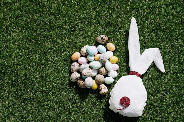 Chocolate Easter eggs glazed with pastel coloring and quail eggs over green grass. Bunny shaped white baking paper. Happy Easter concept. Copy space. Top view