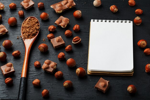 Photo chocolate with hazelnuts, a notebook with blank pages and a wooden spoon with cocoa on a dark background, surrounded by nuts in the shell and peeled.