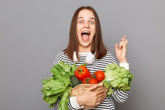 Choose green eat healthy Vitaminrich fruits for wellness Beautyenhancing diet Hopeful excited woman holding vegetables crossed fingers isolated over gray background