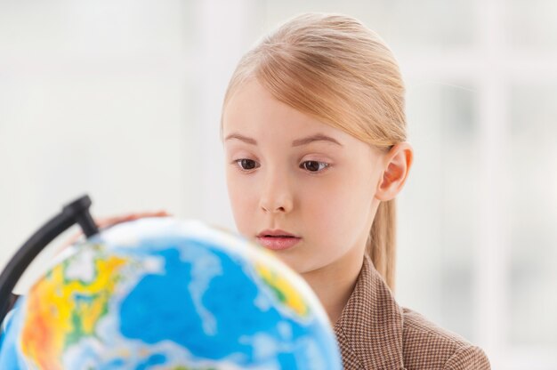 Choosing a place to travel. Concentrated little girl in formalwear examining globe with a loupe while sitting at the table