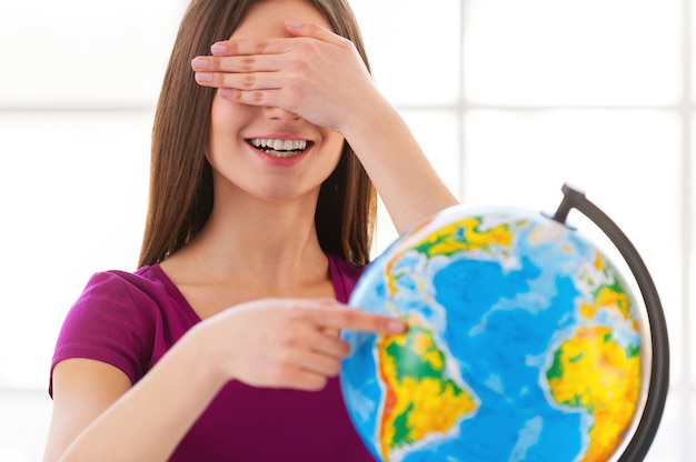 Choosing a traveling point. Cheerful little girl in formalwear examining globe with a loupe while sitting at the table