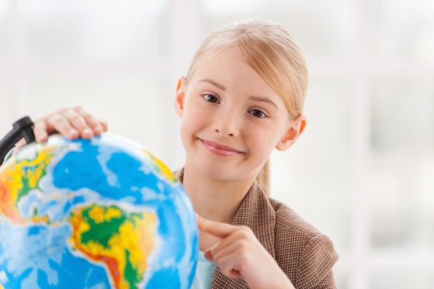 Choosing a traveling point. Cheerful little girl in formalwear examining globe with a loupe while sitting at the table