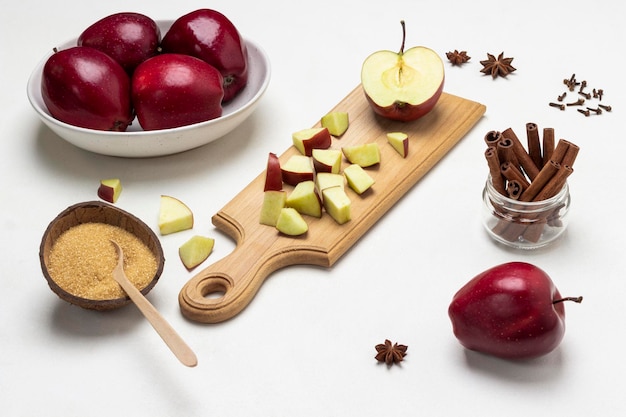 Chopped apples on cutting board Brown sugar in coconut shell Cinnamon sticks in glass jar Ripe red apples in bowl Top view White background