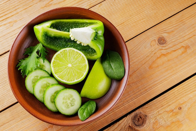 Chopped green fruits and vegetables on a wooden boards. Sliced green apple, bell pepper, cucumber and lime in a clay bowl. Top view. Copy space