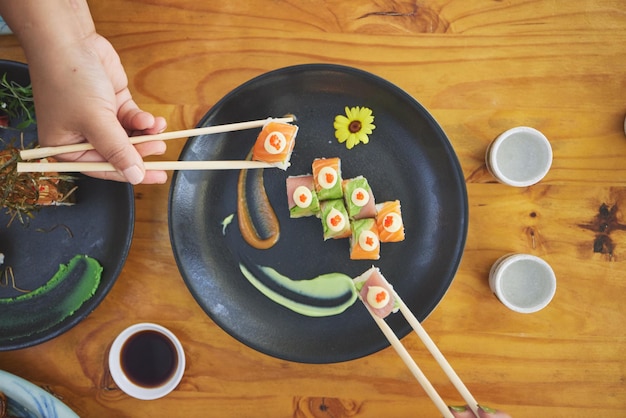 Chopsticks hand and eating sushi and food at a restaurant for nutrition at table Above hungry people with wooden sticks and soy sauce for dining Japanese culture and cuisine with art on a plate