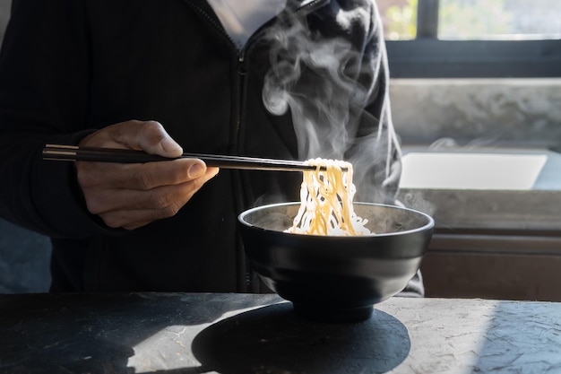 Chopsticks to tasty noodles with steam and smoke in bowl on wooden background, selective focus., Asian meal on a table, junk food concept