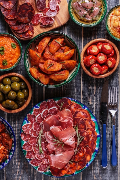 Photo chorizo, salami, bowls with olives,  peppers, anchovies, spicy potatoes, mashed chickpeas on a wooden table.