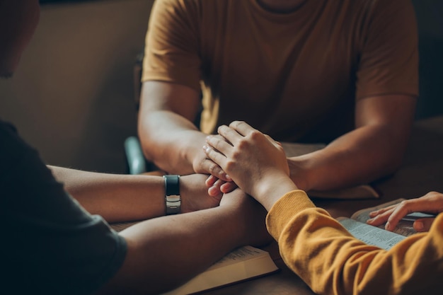 Christian family sitting around a wooden table with open bible page and holding hands to bless