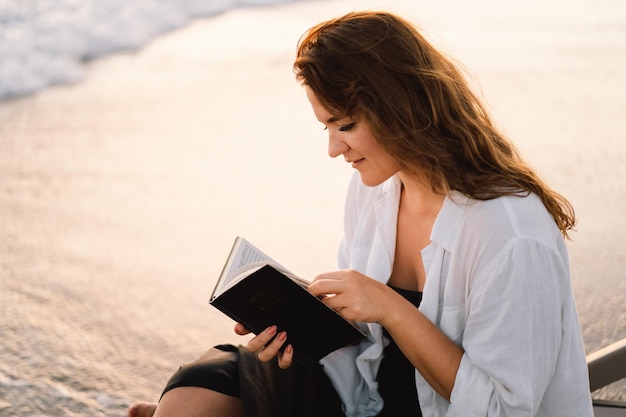 Christian woman holds bible in her hands reading the holy bible on the sea during beautiful sunset