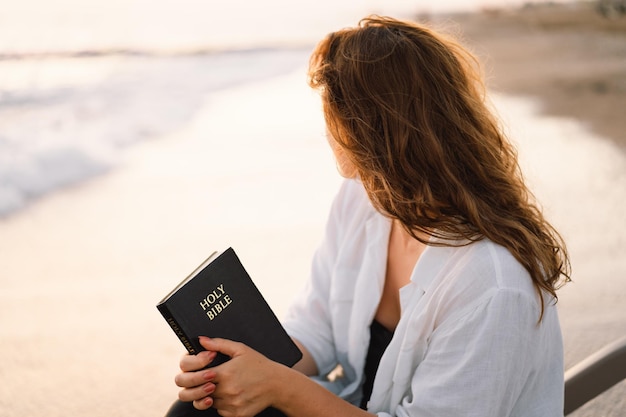 Christian woman holds bible in her hands reading the holy bible on the sea during beautiful sunset