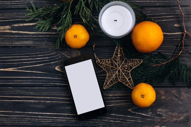 Christmas advertising concept phone with empty screen on christmas background of green branches and oranges and golden star on black rustic wooden desk flat lay space for text